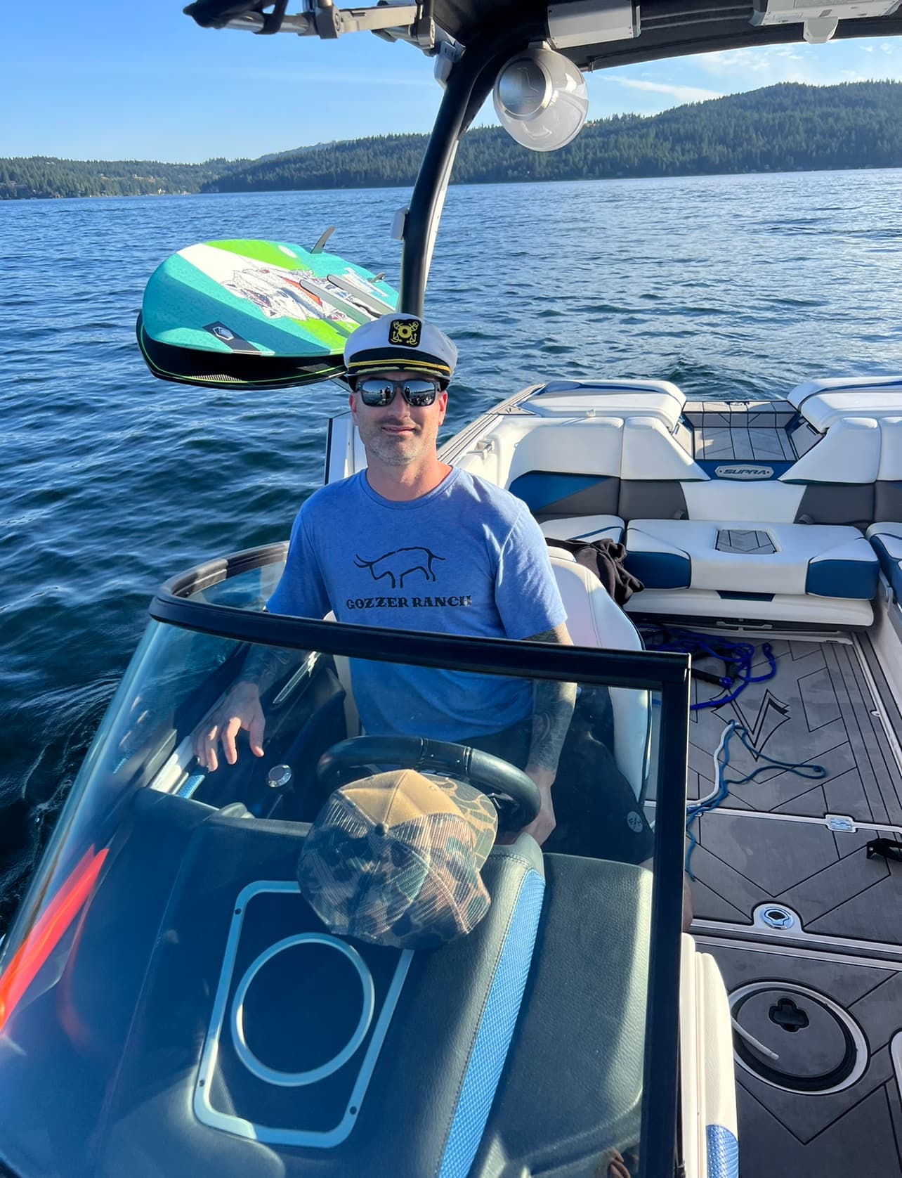 Man in captain's hat and sunglasses at the helm of a boat on a lake.